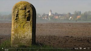 Standbild_aus dem Kurzfilm "Heimatpflege - Kulturlandschaft Bayern digital" vom BR: Sein-Wegmakierung auf dem Acker vor einer Stadt