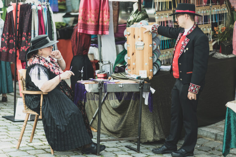 Eine Frau in Tracht sitzt an einem Tisch. Neben Ihr steht ein Mann in Tracht. Sie sind an einem Markt-Stand mit verschiedenen Textilien.