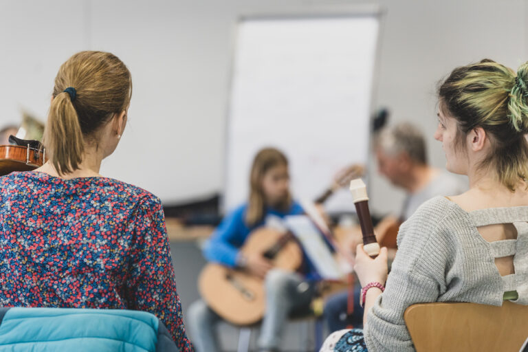 Zwei Frauen von hinten, sitzend, mit Musikinstrumenten in der Hand. Im Hintergrund unscharf mehr Musikanten.