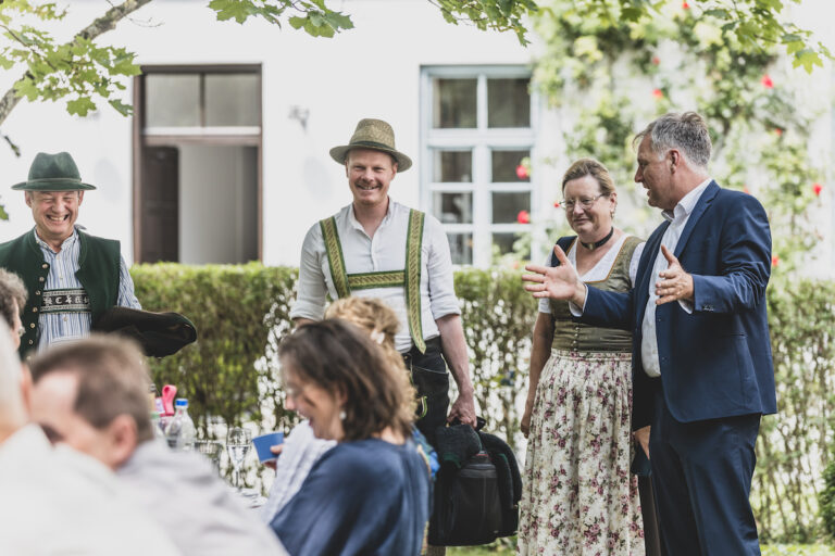 Szene mit mehreren Menschen, davon drei stehend in Tracht und andere an Bierbänken sitzend im Vordergrund in normaler Kleidung. Rudolf Neumaier im Anzug ebenfalls stehend und gestikulierend. Im Hintergrund Fassade des Landesvereins.