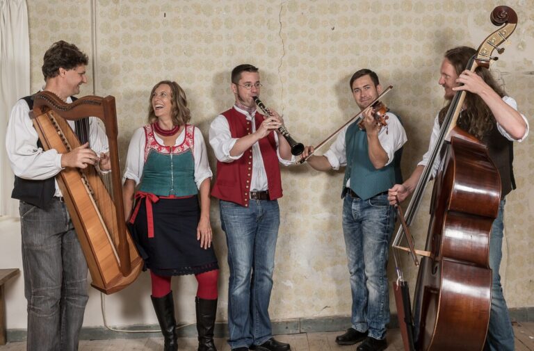 Fünf Personen der Musikgruppe Liadhaber stehen mit ihren Instrumenten nebeneinander in einer Reihe vor einer Wand in einem alten Gebäude