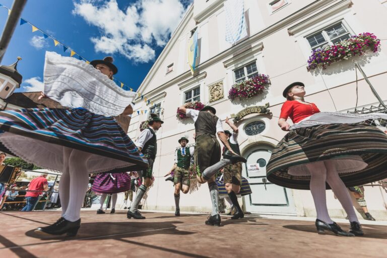Junge Menschen in oberbayerischer Tracht tanzen Schuhplattler auf einer Bühne vor einer prächtigen Rathaus-Fassade bei einem Fest.