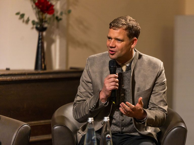 Mann im Trachtenanzug mit Mikrofon auf einem Sessel sitzend, sprechend. Erster Vorsitzender Dr. Olaf Heinrich