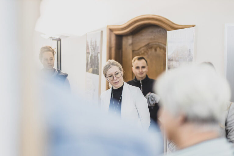 Daniela Sandner vor einer historischen Tür, Blick nach unten rechts. Andere Personen im Vordergrund und Hintergrund unscharf. Erklärtafeln links und rechts neben der Tür.