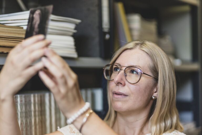 Daniela Sandner hält eine Fotografie-Glasplatte ins Licht. Aufnahme von vorne. Im Hintergrund ein Bücherregal.