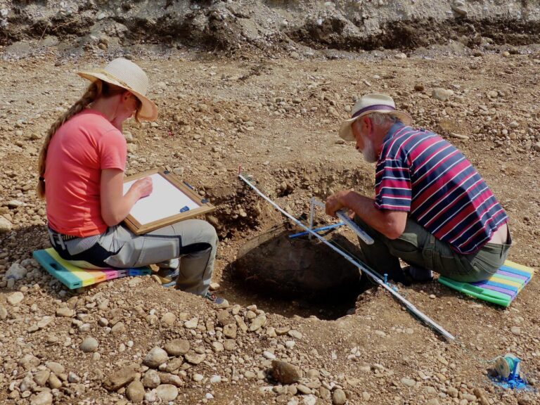 Zwei Archäologen sitzen auf der Erde an einem Loch. Frau links zeichnet, Mann rechts misst im Zollstock.