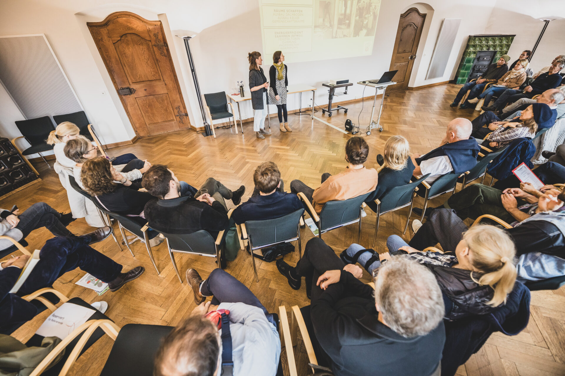 Menschen sitzen in einem Halbkreis auf Stühlen um zwei Frauen, die eine Powerpoint-Präsentation halten. Ansicht von schräg-oben.