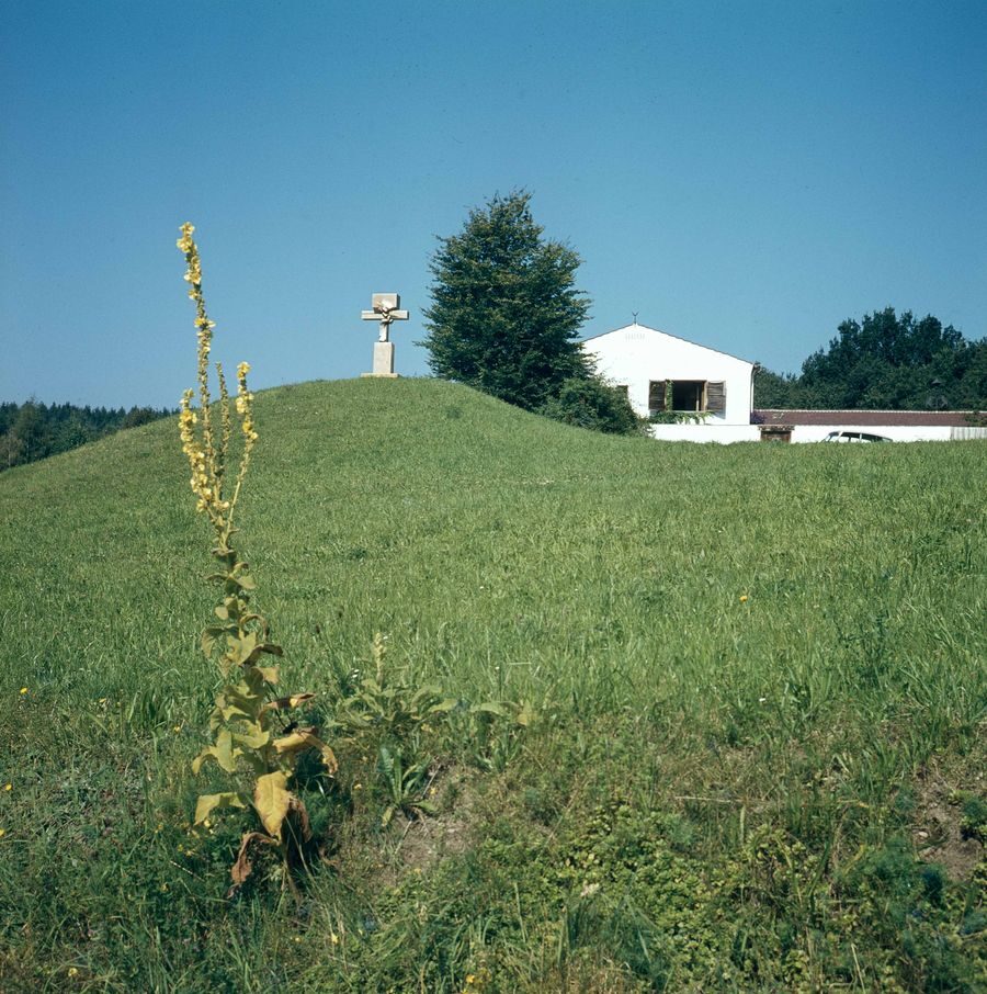 Ein kleines weißes Haus mit Spitzdach auf einem Hügel. Links ein Steinkreuz.