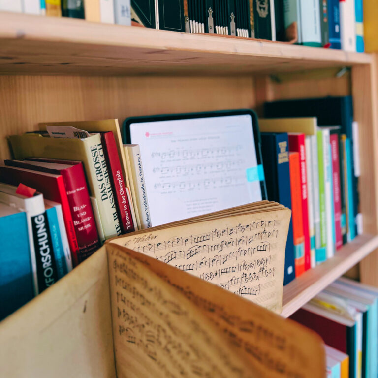 Ein Bücherregal mit einem Tablet darin, auf dem Noten des Landesvereins zus ehen sind. Im Vordergrund ein aufgeschlagenes Notenbuch.