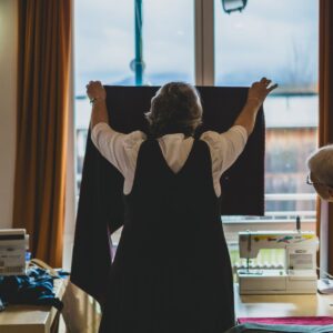 Zwei Frauen arbeiten an einem Kleidungsstück, die eine hält ein Textilstück vor ein Fenster, die andere schaut von der Seite darauf. Nähmaschinen sind auf tischen zu sehen.